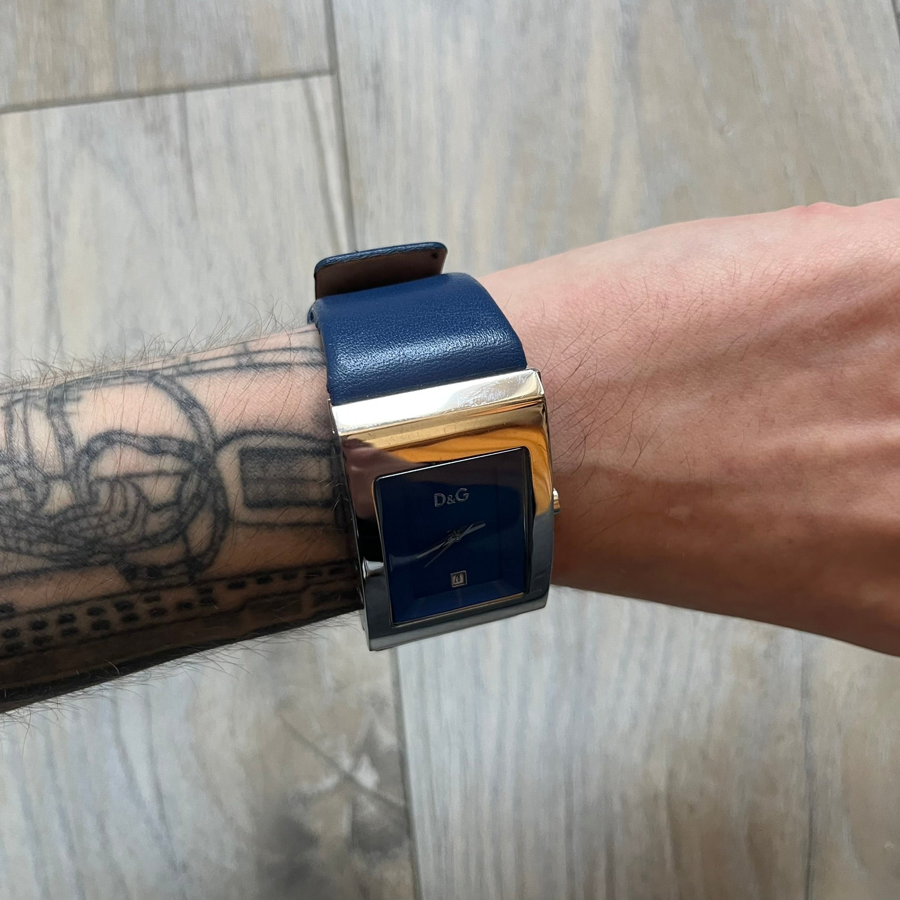 Dolce & Gabbana Navy & Silver Analog Watch features a silver-tone case with a navy dial and matching navy leather strap. The dial has a minimal design with silver hands, a small D&G logo, and a day-date window. The side of the case includes a raised Dolce & Gabbana logo, while the inside of the leather strap is stamped with D&G branding.