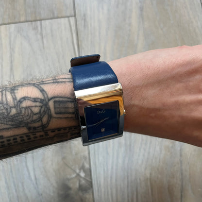 Dolce & Gabbana Navy & Silver Analog Watch features a silver-tone case with a navy dial and matching navy leather strap. The dial has a minimal design with silver hands, a small D&G logo, and a day-date window. The side of the case includes a raised Dolce & Gabbana logo, while the inside of the leather strap is stamped with D&G branding.