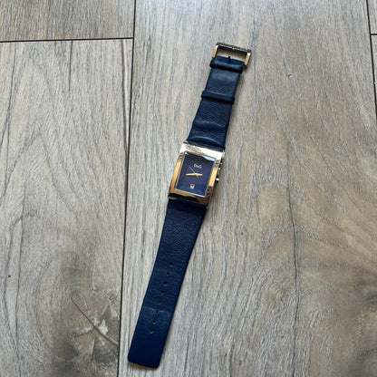 Dolce & Gabbana Navy & Silver Analog Watch features a silver-tone case with a navy dial and matching navy leather strap. The dial has a minimal design with silver hands, a small D&G logo, and a day-date window. The side of the case includes a raised Dolce & Gabbana logo, while the inside of the leather strap is stamped with D&G branding.