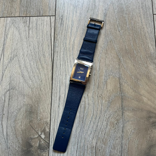 Dolce & Gabbana Navy & Silver Analog Watch features a silver-tone case with a navy dial and matching navy leather strap. The dial has a minimal design with silver hands, a small D&G logo, and a day-date window. The side of the case includes a raised Dolce & Gabbana logo, while the inside of the leather strap is stamped with D&G branding.