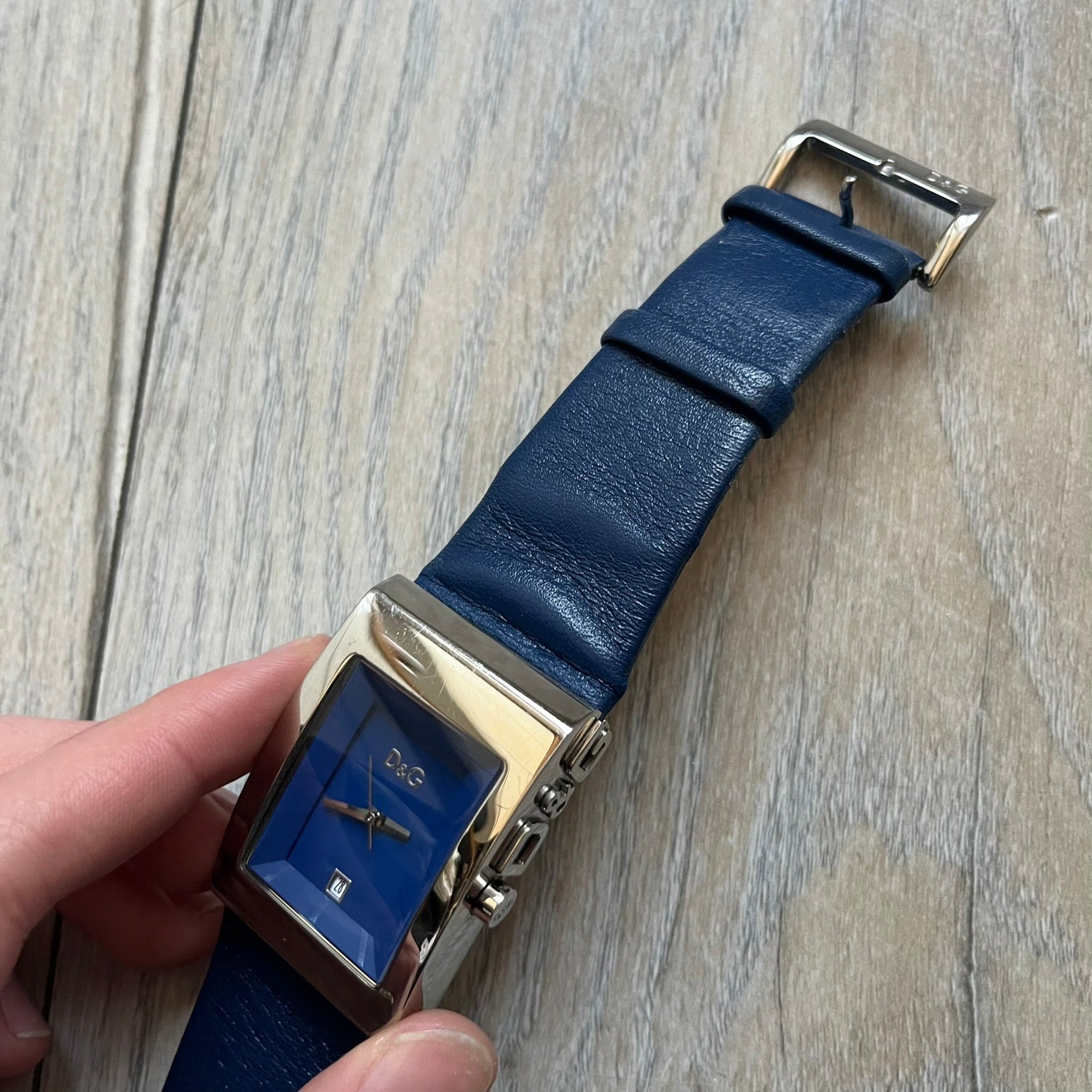 Dolce & Gabbana Navy & Silver Analog Watch features a silver-tone case with a navy dial and matching navy leather strap. The dial has a minimal design with silver hands, a small D&G logo, and a day-date window. The side of the case includes a raised Dolce & Gabbana logo, while the inside of the leather strap is stamped with D&G branding.