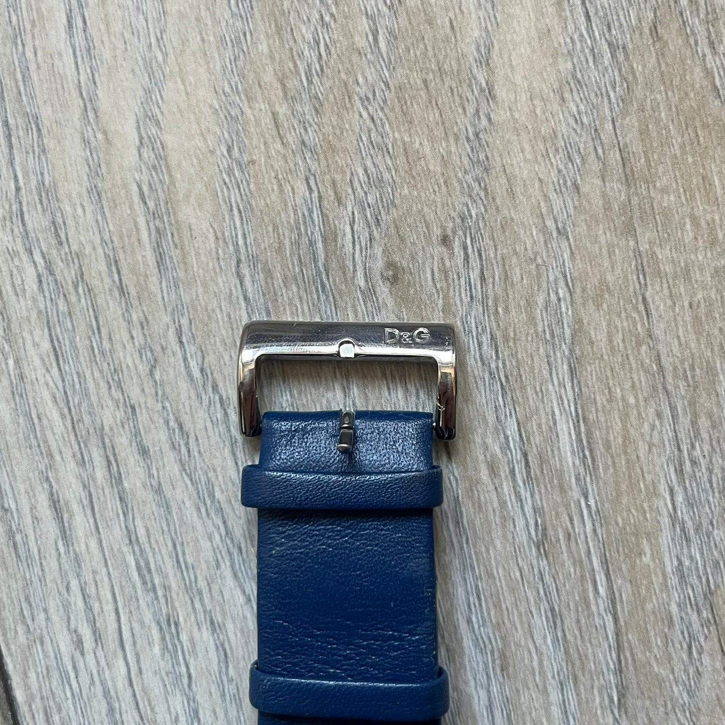 Dolce & Gabbana Navy & Silver Analog Watch features a silver-tone case with a navy dial and matching navy leather strap. The dial has a minimal design with silver hands, a small D&G logo, and a day-date window. The side of the case includes a raised Dolce & Gabbana logo, while the inside of the leather strap is stamped with D&G branding.