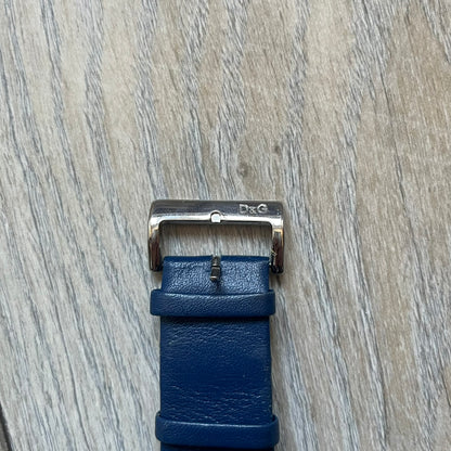 Dolce & Gabbana Navy & Silver Analog Watch features a silver-tone case with a navy dial and matching navy leather strap. The dial has a minimal design with silver hands, a small D&G logo, and a day-date window. The side of the case includes a raised Dolce & Gabbana logo, while the inside of the leather strap is stamped with D&G branding.