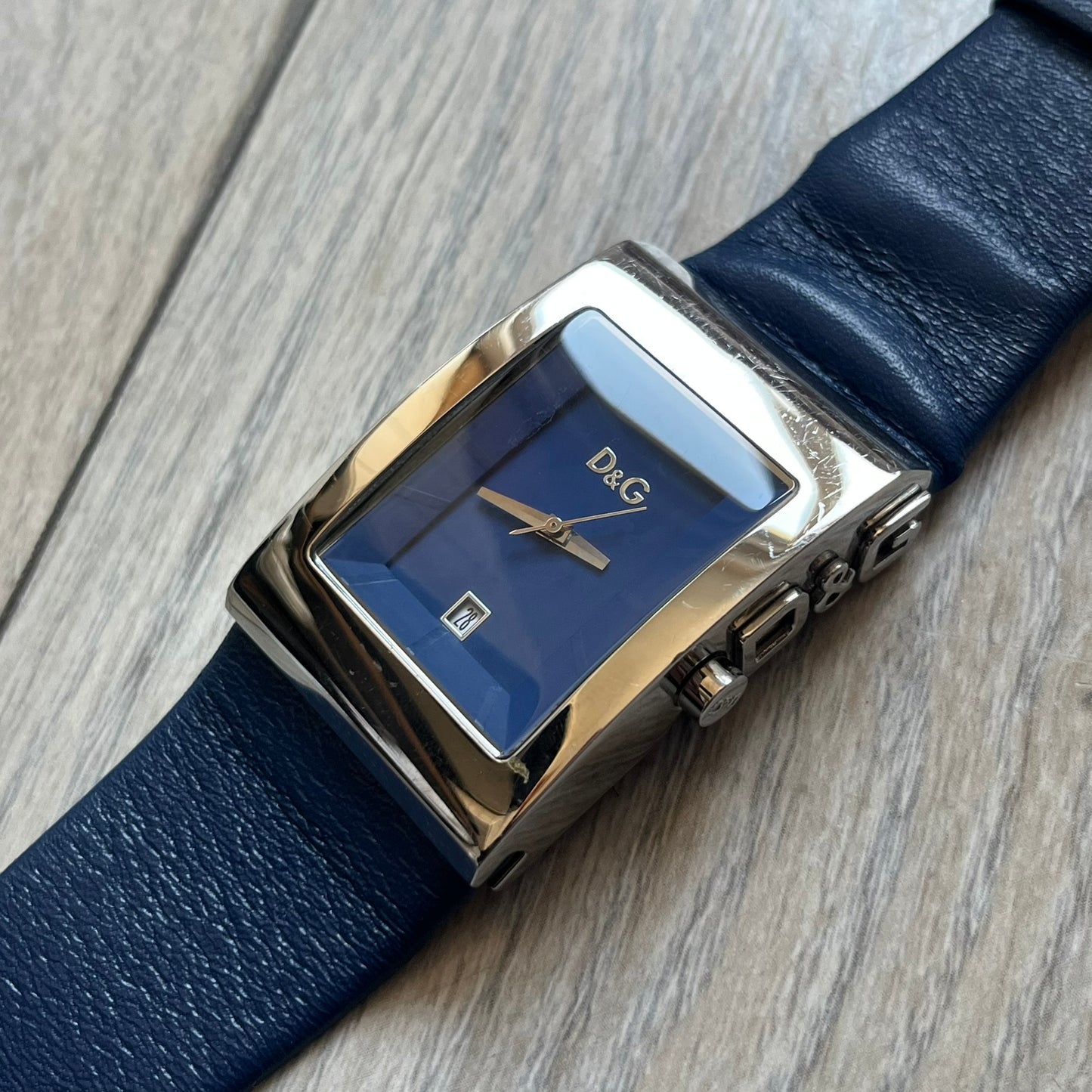 Dolce & Gabbana Navy & Silver Analog Watch features a silver-tone case with a navy dial and matching navy leather strap. The dial has a minimal design with silver hands, a small D&G logo, and a day-date window. The side of the case includes a raised Dolce & Gabbana logo, while the inside of the leather strap is stamped with D&G branding.