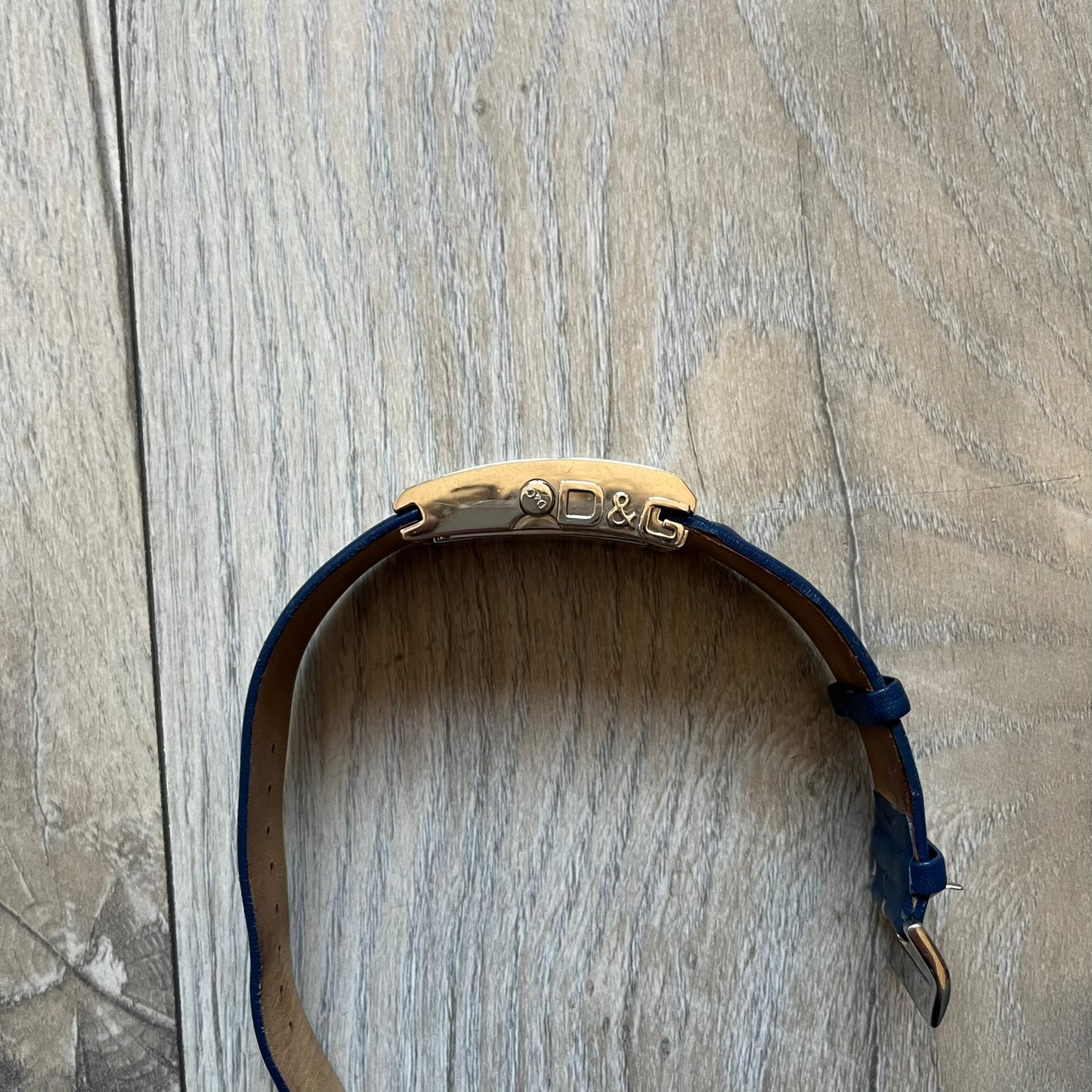 Dolce & Gabbana Navy & Silver Analog Watch features a silver-tone case with a navy dial and matching navy leather strap. The dial has a minimal design with silver hands, a small D&G logo, and a day-date window. The side of the case includes a raised Dolce & Gabbana logo, while the inside of the leather strap is stamped with D&G branding.