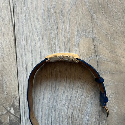 Dolce & Gabbana Navy & Silver Analog Watch features a silver-tone case with a navy dial and matching navy leather strap. The dial has a minimal design with silver hands, a small D&G logo, and a day-date window. The side of the case includes a raised Dolce & Gabbana logo, while the inside of the leather strap is stamped with D&G branding.