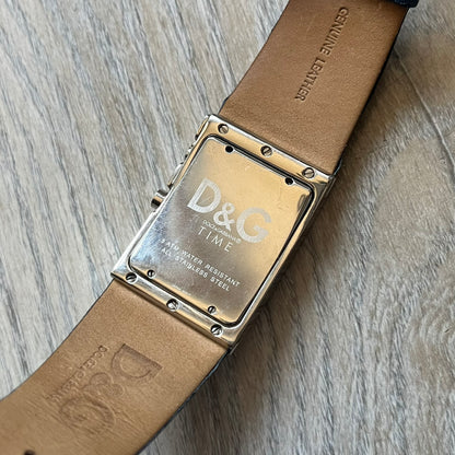 Dolce & Gabbana Navy & Silver Analog Watch features a silver-tone case with a navy dial and matching navy leather strap. The dial has a minimal design with silver hands, a small D&G logo, and a day-date window. The side of the case includes a raised Dolce & Gabbana logo, while the inside of the leather strap is stamped with D&G branding.