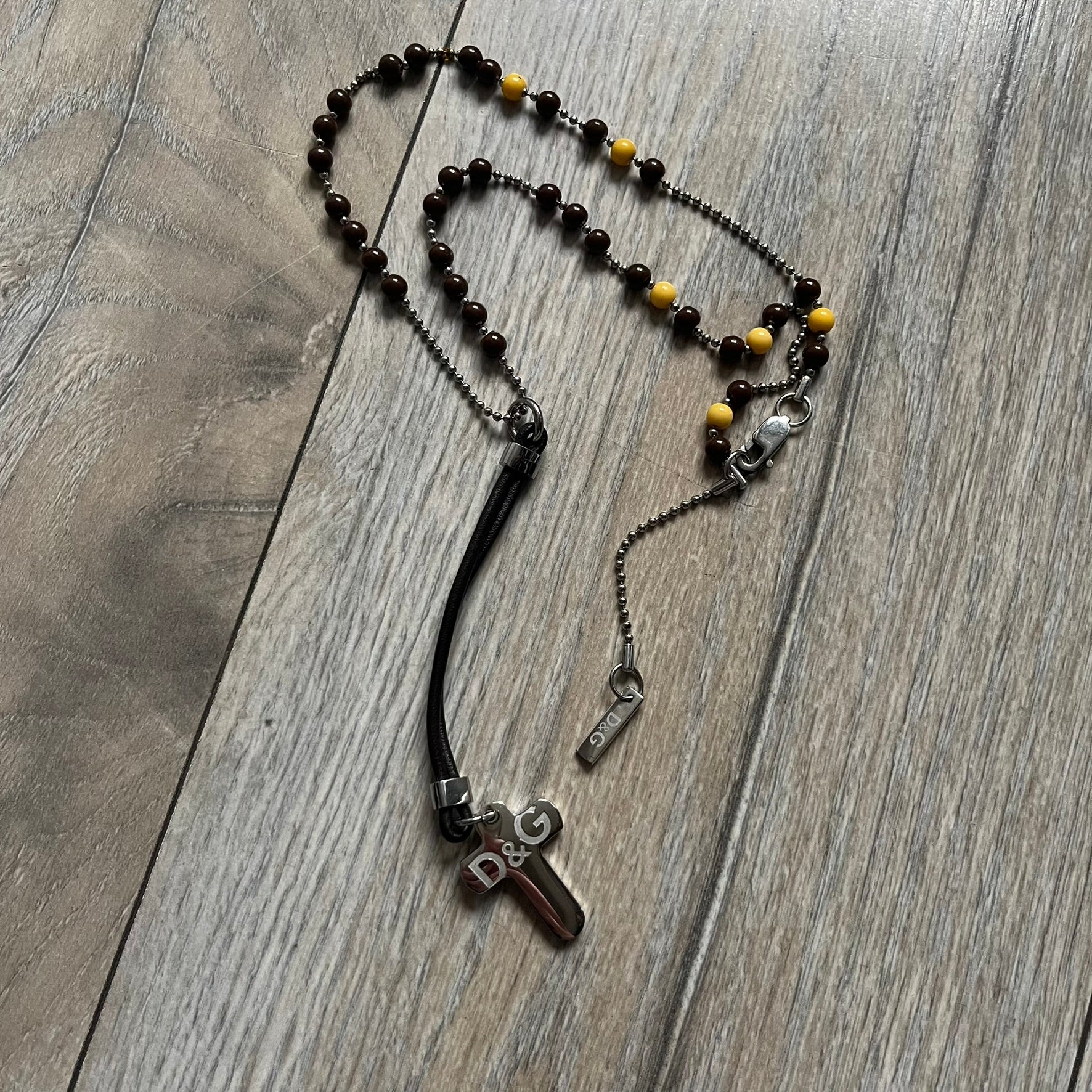 Dolce &amp; Gabbana Pearl Brown Rosary Necklace from the 2000s era. The necklace features a silver-tone metal cross with a D&amp;G logo, connected via a black leather link to a metal chain with alternating brown and yellow pearl-style beads. Rosary-style construction with mixed materials including metal, leather, and resin pearls. Comes complete with the big dustbag.