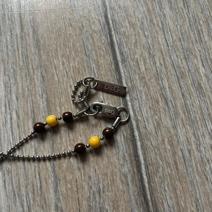 Dolce &amp; Gabbana Pearl Brown Rosary Necklace from the 2000s era. The necklace features a silver-tone metal cross with a D&amp;G logo, connected via a black leather link to a metal chain with alternating brown and yellow pearl-style beads. Rosary-style construction with mixed materials including metal, leather, and resin pearls. Comes complete with the big dustbag.