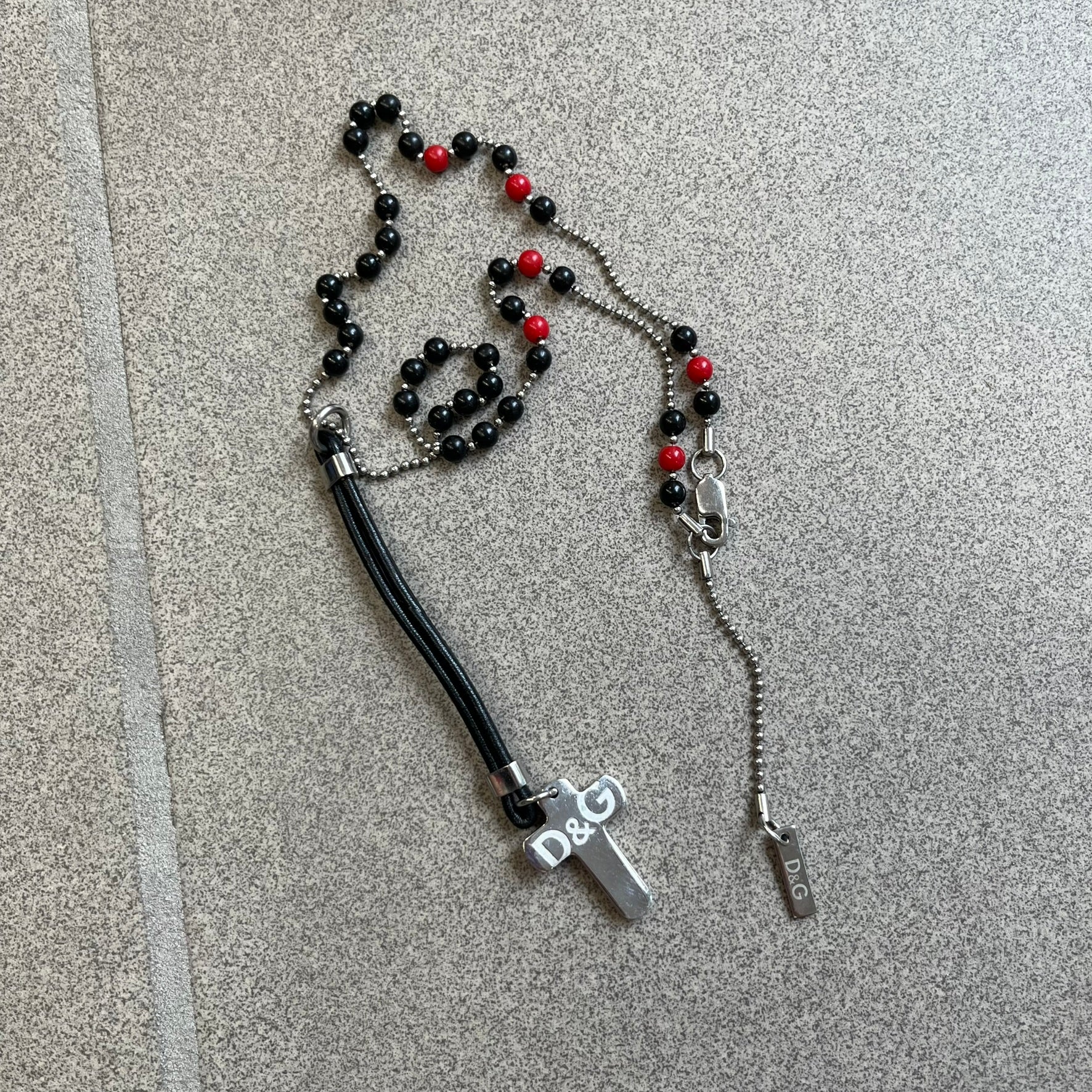 Dolce &amp; Gabbana Pearl Red/Black Rosary Necklace from the 2000's era. The necklace features a silver-tone metal cross with a D&amp;G logo, connected via a black leather link to a metal chain with alternating red and black pearl-style beads. Rosary-style construction with mixed materials including metal, leather, and resin pearls.&nbsp;