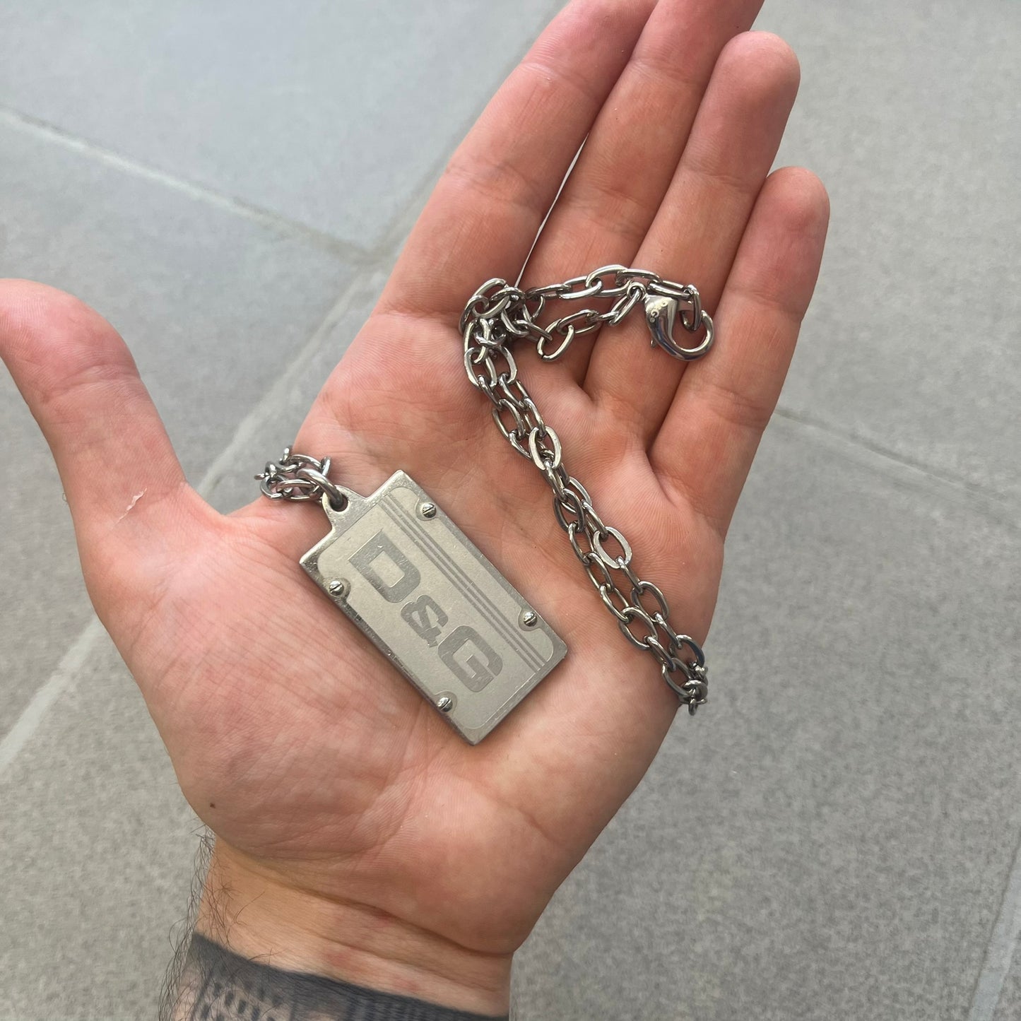 Dolce &amp; Gabbana Propaganda Dog Tag Necklace, featuring a silver stainless steel square tag with four visible screws and the D&amp;G logo engraved on the front. The tag hangs from a durable cable chain, combining sturdy construction with clean, industrial-inspired detailing.