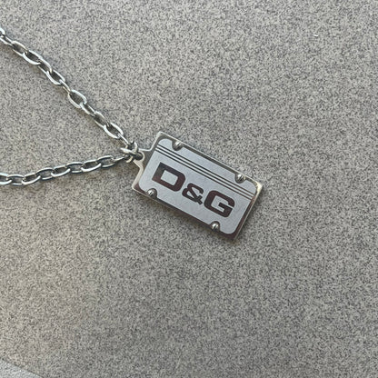 Dolce &amp; Gabbana Propaganda Dog Tag Necklace, featuring a silver stainless steel square tag with four visible screws and the D&amp;G logo engraved on the front. The tag hangs from a durable cable chain, combining sturdy construction with clean, industrial-inspired detailing.