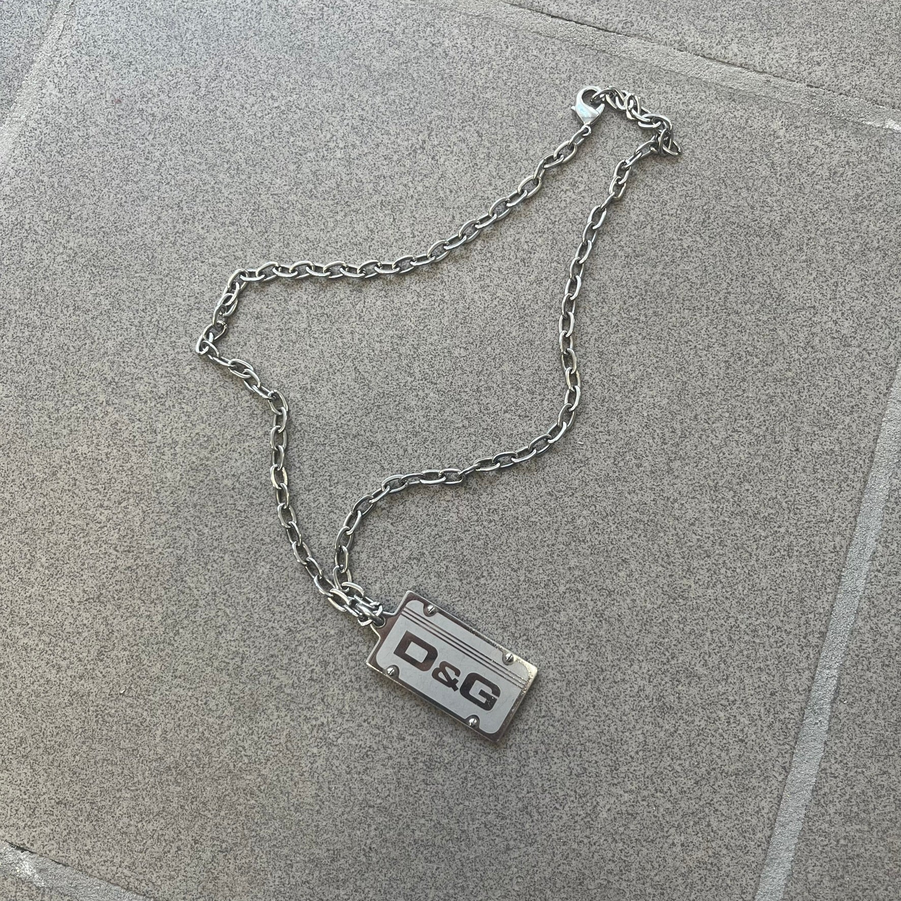 Dolce &amp; Gabbana Propaganda Dog Tag Necklace, featuring a silver stainless steel square tag with four visible screws and the D&amp;G logo engraved on the front. The tag hangs from a durable cable chain, combining sturdy construction with clean, industrial-inspired detailing.