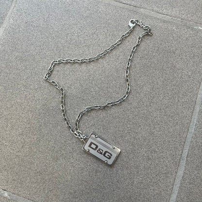 Dolce &amp; Gabbana Propaganda Dog Tag Necklace, featuring a silver stainless steel square tag with four visible screws and the D&amp;G logo engraved on the front. The tag hangs from a durable cable chain, combining sturdy construction with clean, industrial-inspired detailing.