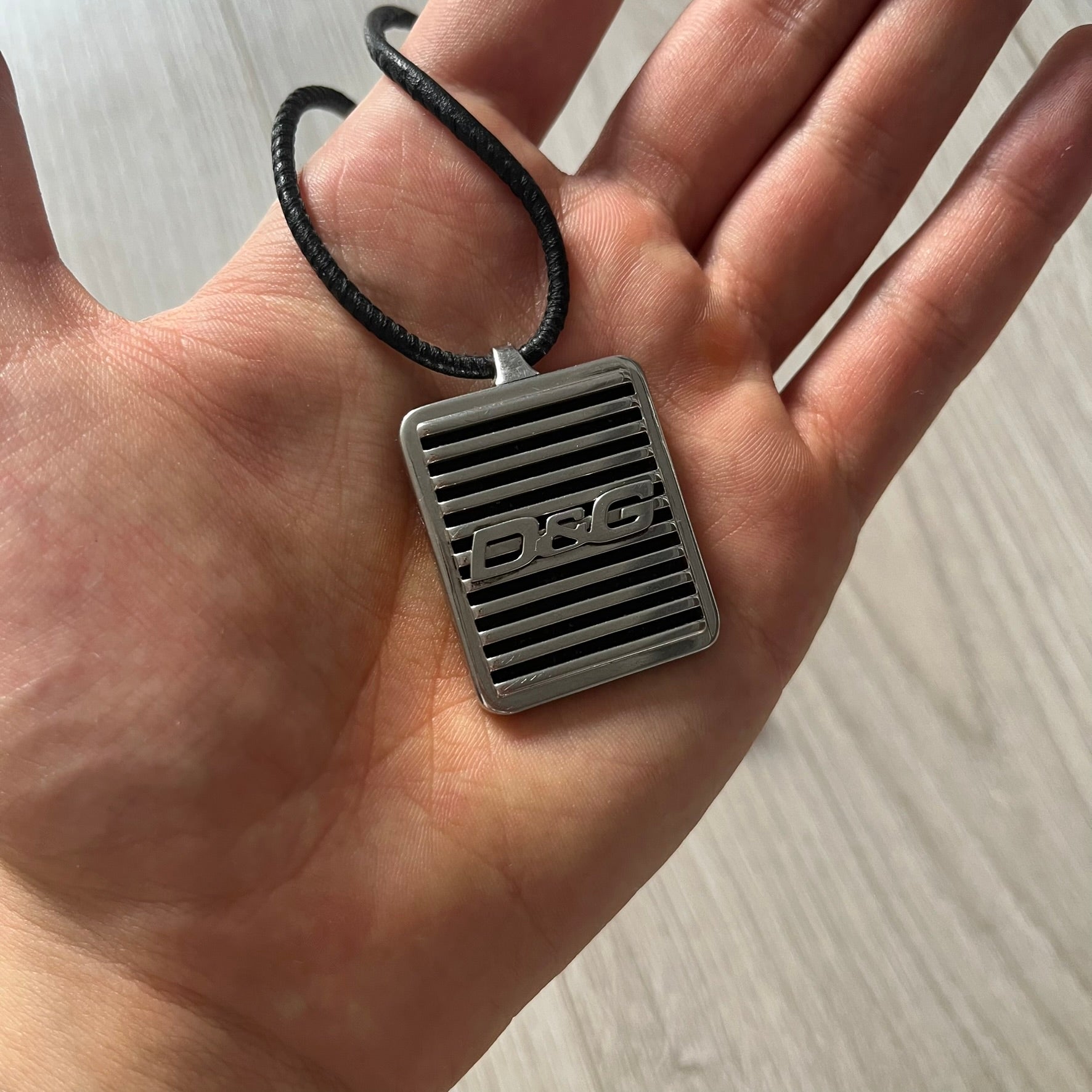 Silver pendant with 'DG' logo held in a hand against a neutral background