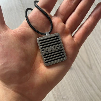 Silver pendant with 'DG' logo held in a hand against a neutral background