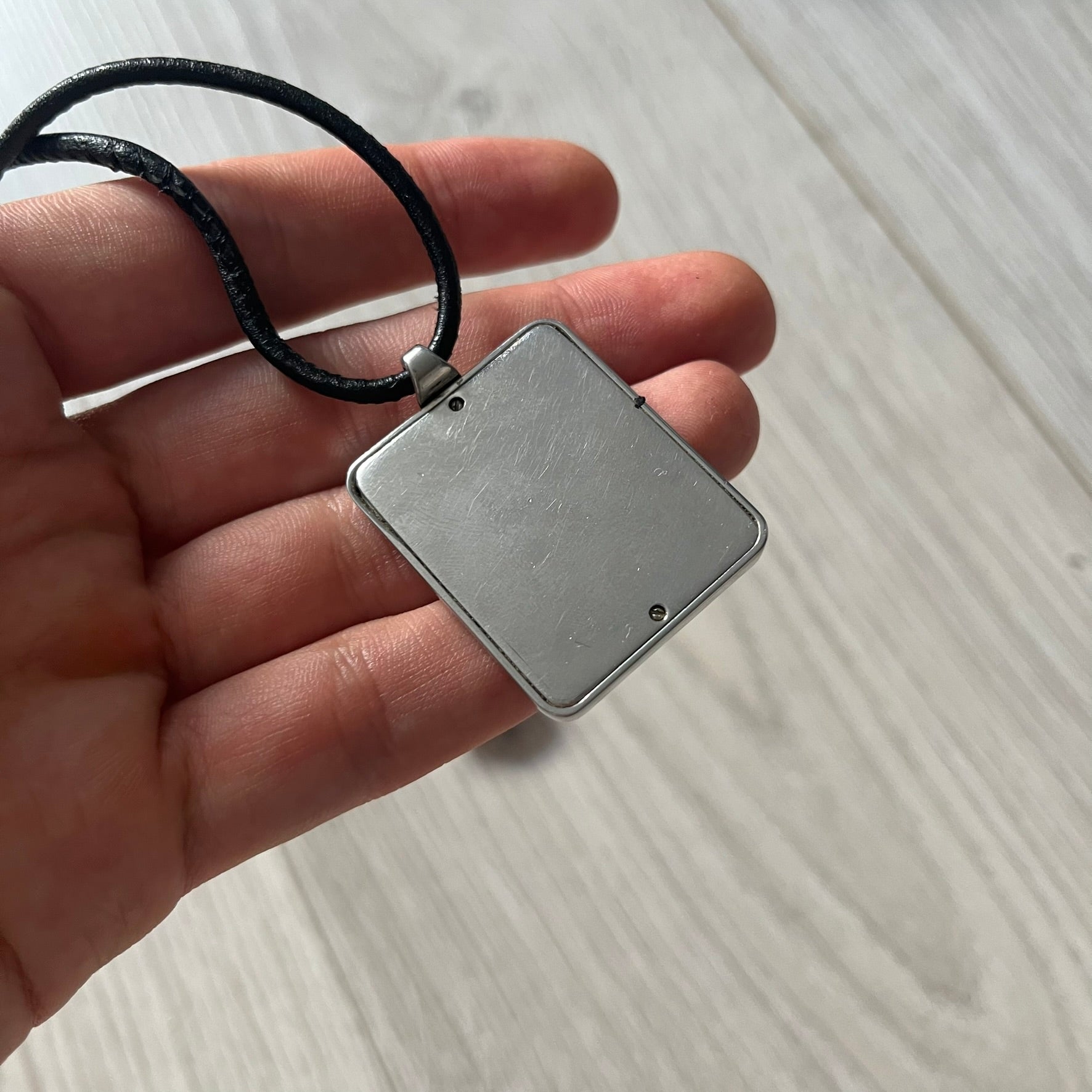 Hand holding a square silver pendant with a black cord against a light wooden surface