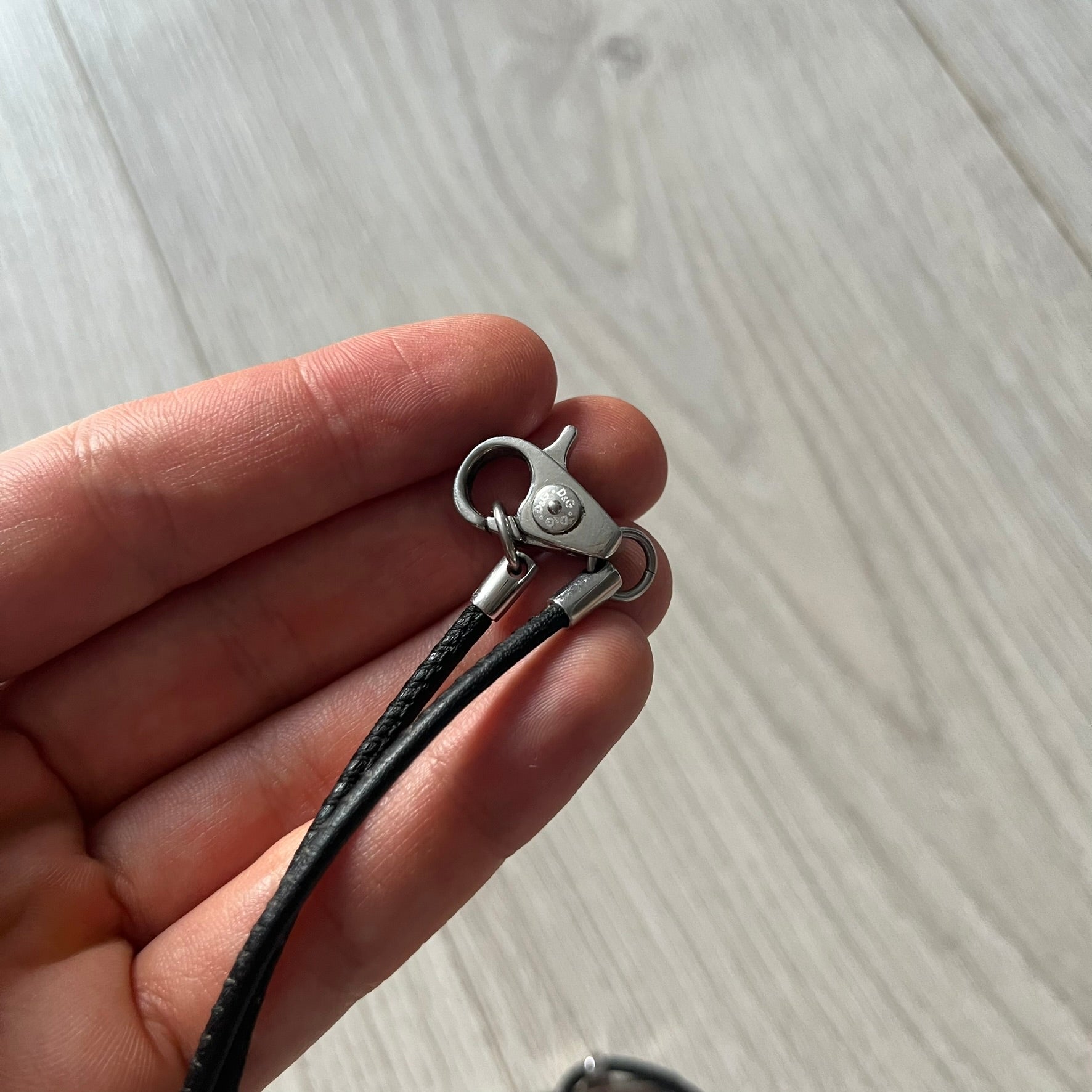Hand holding a small metal clasp with black cord against a light wooden surface