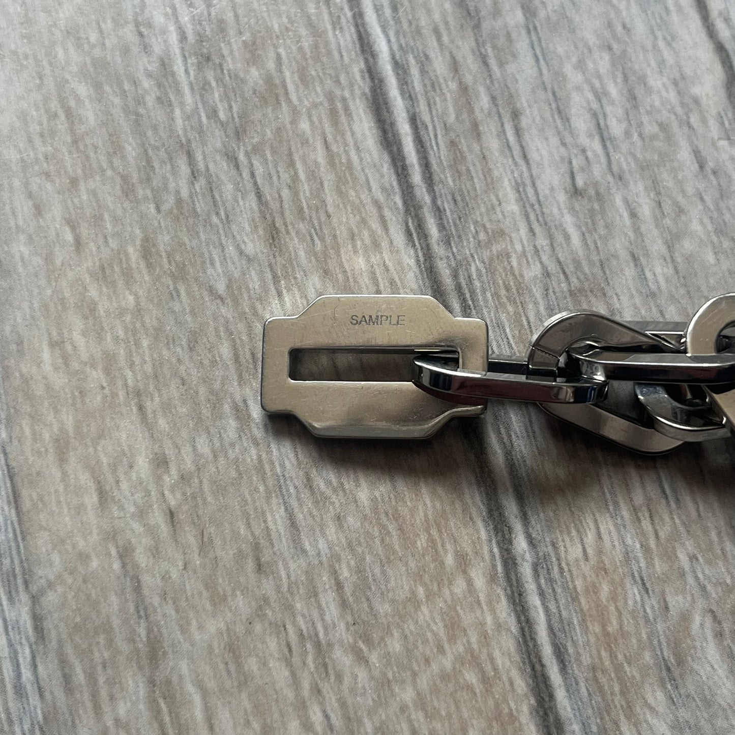 Dolce &amp; Gabbana Razorblade Sample Bracelet is a stainless steel chain bracelet with large interlocking links and a central razor blade-shaped pendant matching the link size. One side of the pendant is engraved with the D&amp;G logo, while the opposite side features small “SAMPLE” text. The bracelet secures with a lobster clasp engraved with Dolce &amp; Gabbana logos.
