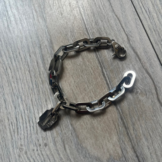 Dolce &amp; Gabbana Razorblade Sample Bracelet is a stainless steel chain bracelet with large interlocking links and a central razor blade-shaped pendant matching the link size. One side of the pendant is engraved with the D&amp;G logo, while the opposite side features small “SAMPLE” text. The bracelet secures with a lobster clasp engraved with Dolce &amp; Gabbana logos.