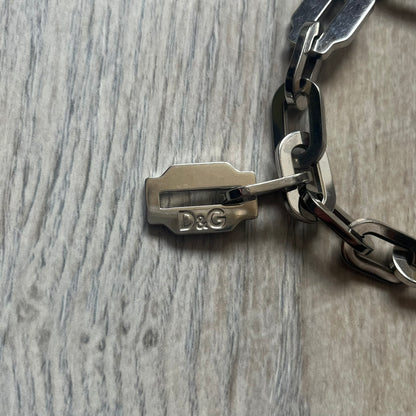 Dolce &amp; Gabbana Razorblade Sample Bracelet is a stainless steel chain bracelet with large interlocking links and a central razor blade-shaped pendant matching the link size. One side of the pendant is engraved with the D&amp;G logo, while the opposite side features small “SAMPLE” text. The bracelet secures with a lobster clasp engraved with Dolce &amp; Gabbana logos.