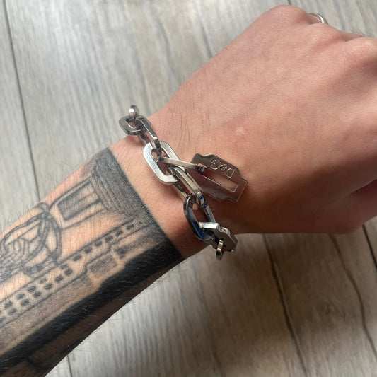 Dolce &amp; Gabbana Razorblade Sample Bracelet is a stainless steel chain bracelet with large interlocking links and a central razor blade-shaped pendant matching the link size. One side of the pendant is engraved with the D&amp;G logo, while the opposite side features small “SAMPLE” text. The bracelet secures with a lobster clasp engraved with Dolce &amp; Gabbana logos.