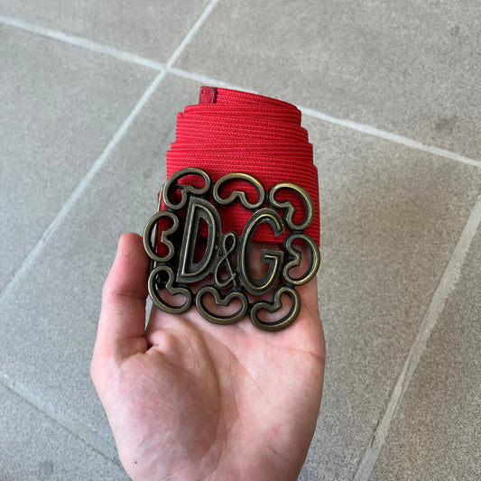 Dolce &amp; Gabbana Red Elastic Hearts Belt is made from red elastic material. The buckle is a metal D&amp;G logo surrounded by eight heart shapes pointing outward, all finished in a dark, aged-looking metal tone. The belt comes with the original hologram tag attached.