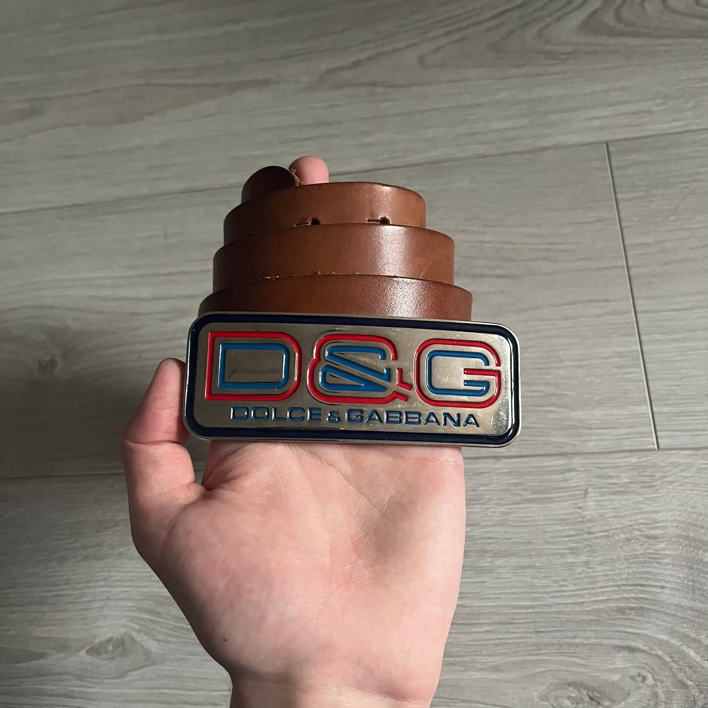 Dolce &amp; Gabbana Red &amp; Blue Leather Belt featuring a rectangular silver-tone buckle with a navy border. The buckle displays a large D&amp;G logo in blue with a red outline and “Dolce &amp; Gabbana” text in blue beneath. Finished with a brown leather strap.