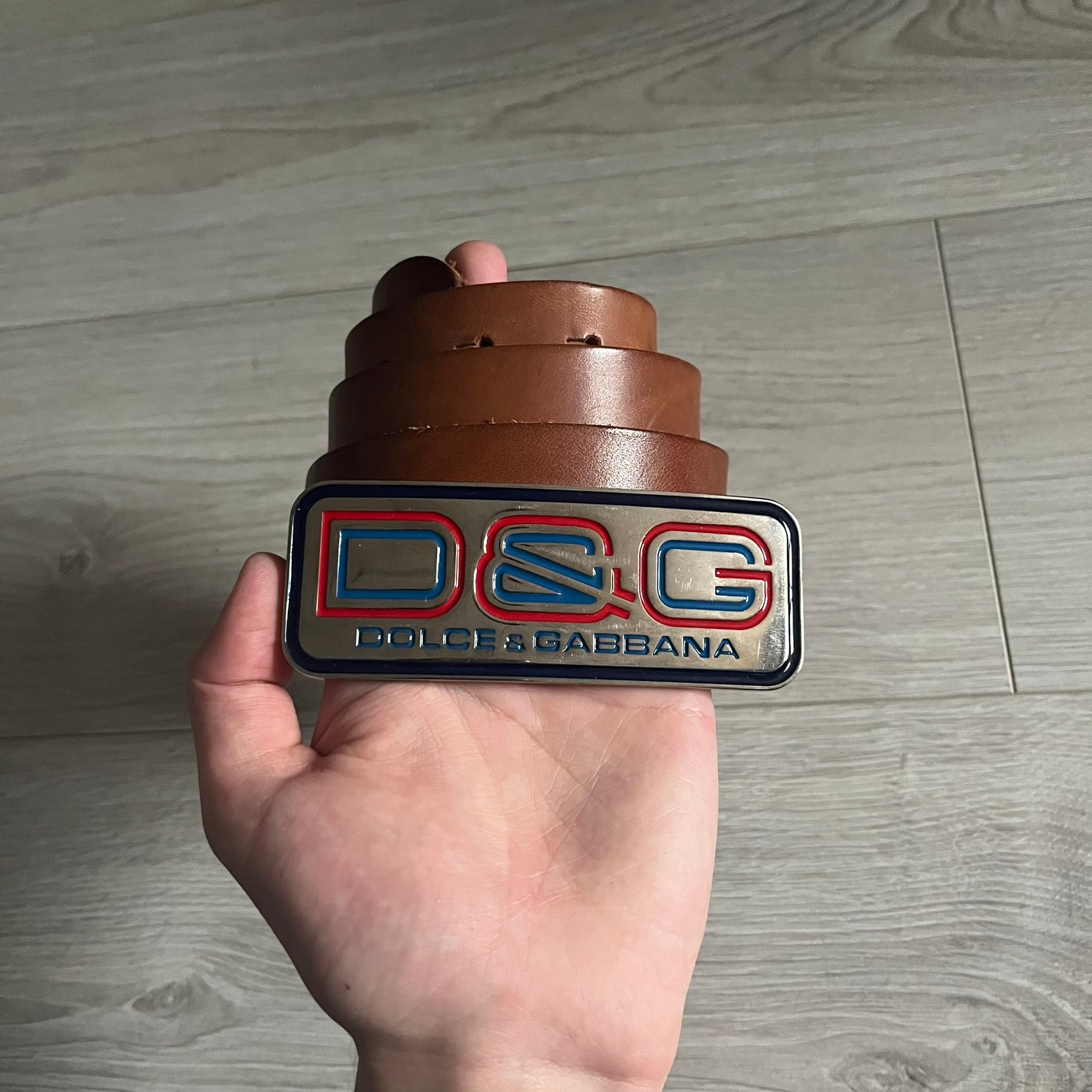 Dolce &amp; Gabbana Red &amp; Blue Leather Belt featuring a rectangular silver-tone buckle with a navy border. The buckle displays a large D&amp;G logo in blue with a red outline and “Dolce &amp; Gabbana” text in blue beneath. Finished with a brown leather strap.