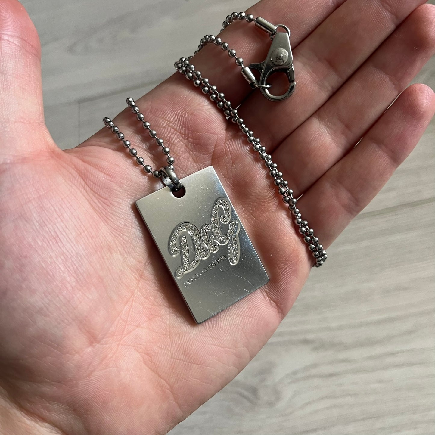 Dolce & Gabbana Rhinestones Dog Tag Necklace DJ0704 crafted from polished stainless steel. Features a rectangular pendant with the iconic D&G logo detailed in rhinestones, and “Dolce & Gabbana” engraved subtly below. Secured with a lobster clasp and finished with a durable ball chain.