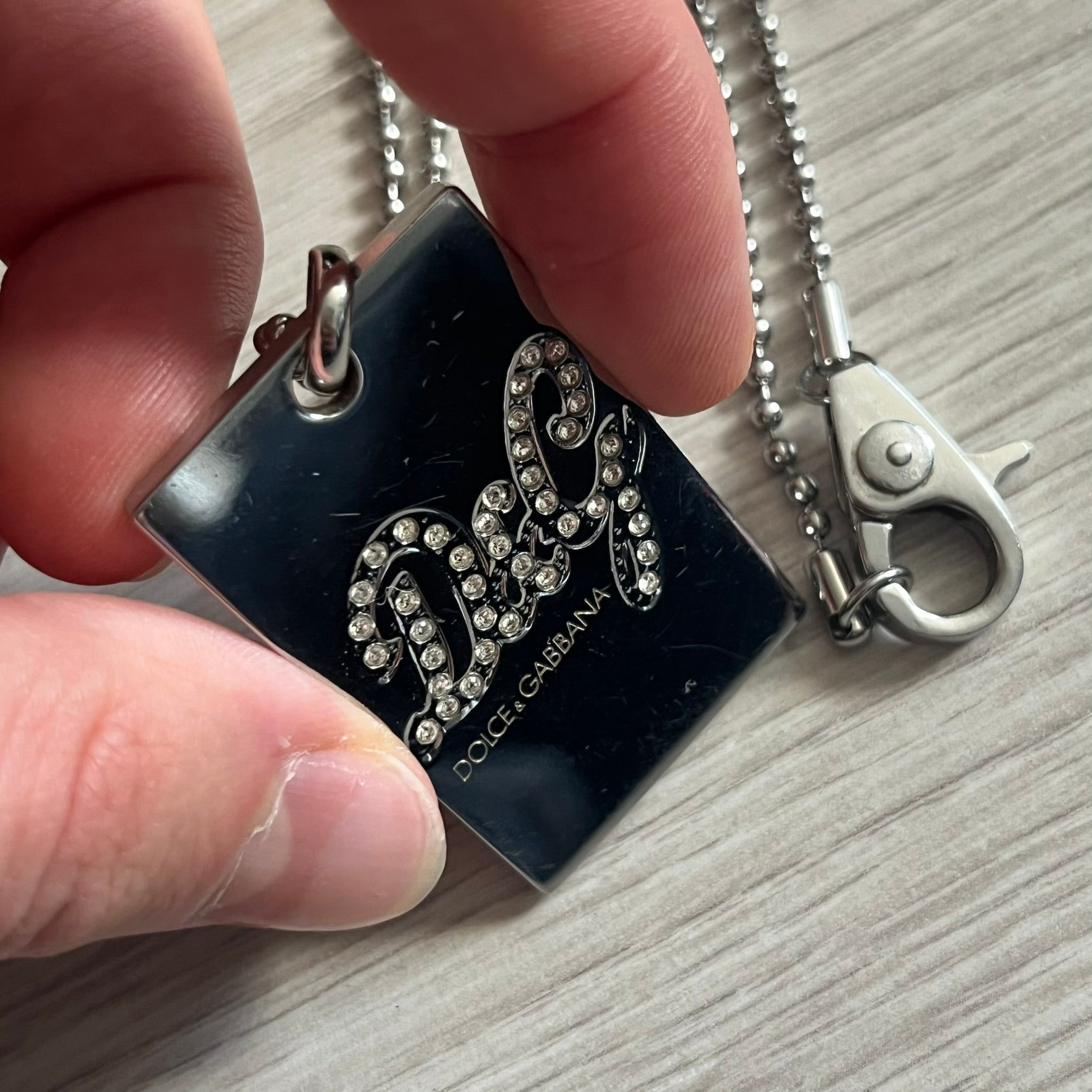 Dolce & Gabbana Rhinestones Dog Tag Necklace DJ0704 crafted from polished stainless steel. Features a rectangular pendant with the iconic D&G logo detailed in rhinestones, and “Dolce & Gabbana” engraved subtly below. Secured with a lobster clasp and finished with a durable ball chain.