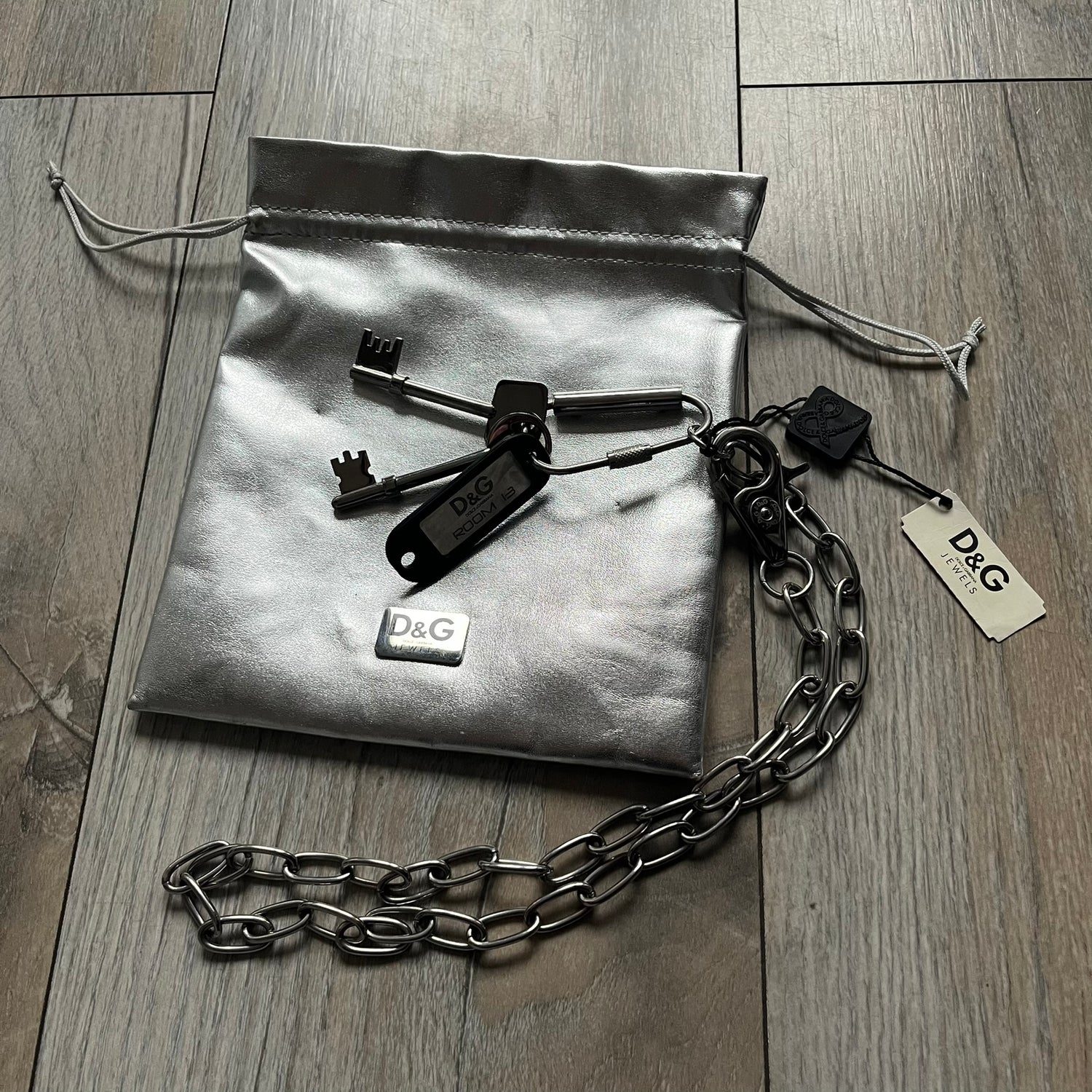 The Dolce &amp; Gabbana Room 13 Carabiner Necklace (DJ0613) is a large, heavy silver-tone necklace featuring a thick paperclip chain with oversized links. At the end of the chain is a large lobster clasp engraved with D&amp;G markings, connected to a branded carabiner. Hanging from the carabiner are two different keys with small D&amp;G logos and a black key identifier with a silver-tone plate engraved with “D&amp;G Dolce &amp; Gabbana Room 13.” The necklace comes with a large silver Dolce &amp; Gabbana dus