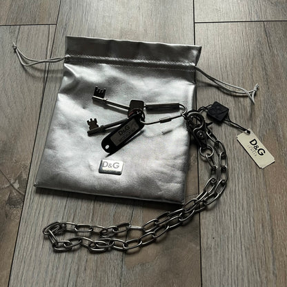 The Dolce &amp; Gabbana Room 13 Carabiner Necklace (DJ0613) is a large, heavy silver-tone necklace featuring a thick paperclip chain with oversized links. At the end of the chain is a large lobster clasp engraved with D&amp;G markings, connected to a branded carabiner. Hanging from the carabiner are two different keys with small D&amp;G logos and a black key identifier with a silver-tone plate engraved with “D&amp;G Dolce &amp; Gabbana Room 13.” The necklace comes with a large silver Dolce &amp; Gabbana dus