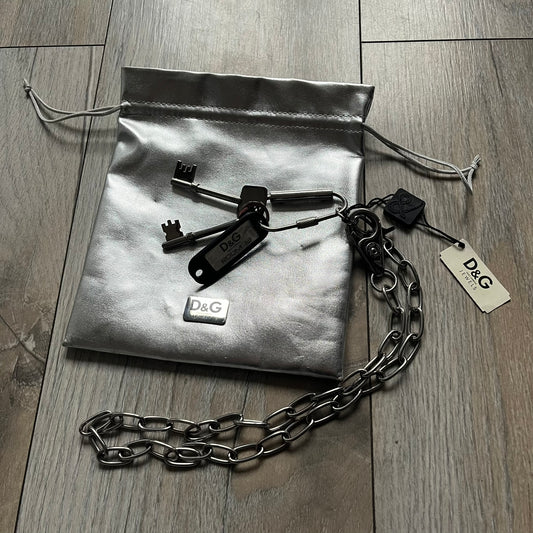 The Dolce &amp; Gabbana Room 13 Carabiner Necklace (DJ0613) is a large, heavy silver-tone necklace featuring a thick paperclip chain with oversized links. At the end of the chain is a large lobster clasp engraved with D&amp;G markings, connected to a branded carabiner. Hanging from the carabiner are two different keys with small D&amp;G logos and a black key identifier with a silver-tone plate engraved with “D&amp;G Dolce &amp; Gabbana Room 13.” The necklace comes with a large silver Dolce &amp; Gabbana dus