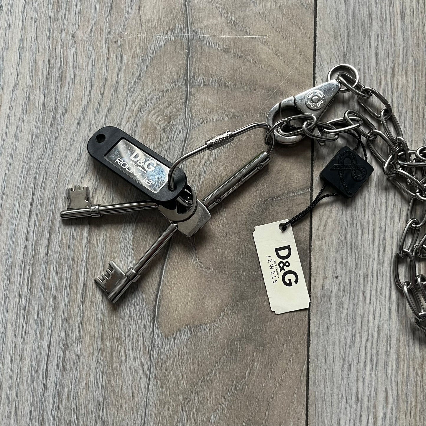 The Dolce &amp; Gabbana Room 13 Carabiner Necklace (DJ0613) is a large, heavy silver-tone necklace featuring a thick paperclip chain with oversized links. At the end of the chain is a large lobster clasp engraved with D&amp;G markings, connected to a branded carabiner. Hanging from the carabiner are two different keys with small D&amp;G logos and a black key identifier with a silver-tone plate engraved with “D&amp;G Dolce &amp; Gabbana Room 13.” The necklace comes with a large silver Dolce &amp; Gabbana dus