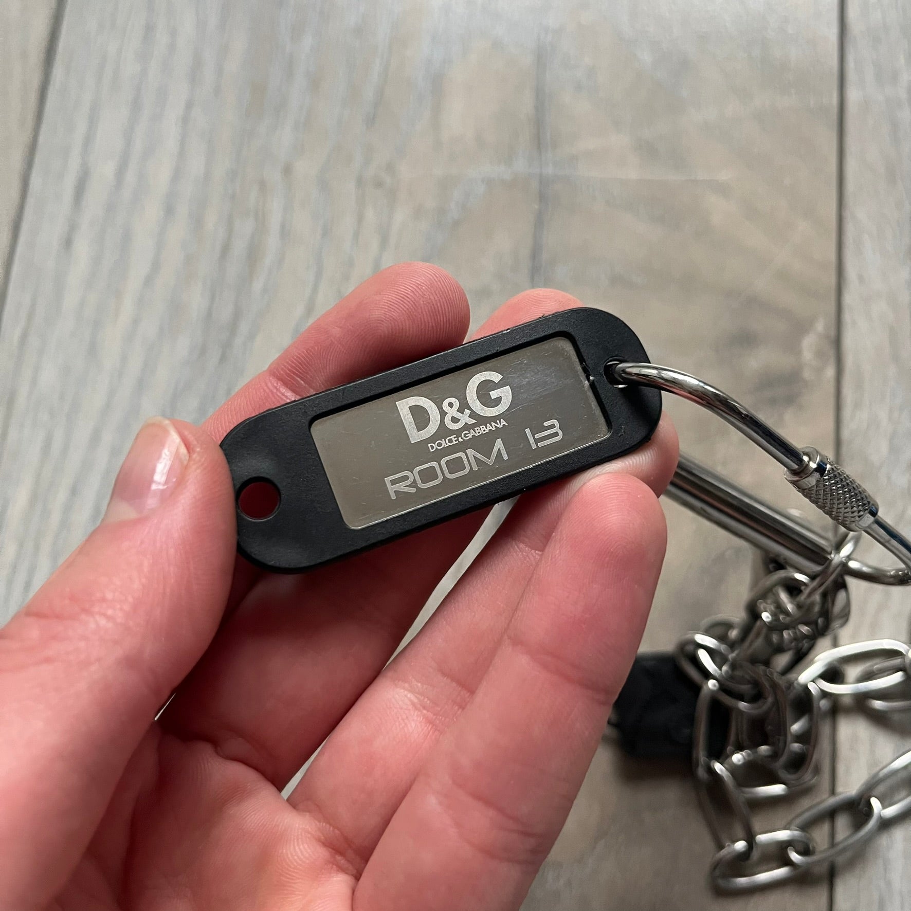 The Dolce &amp; Gabbana Room 13 Carabiner Necklace (DJ0613) is a large, heavy silver-tone necklace featuring a thick paperclip chain with oversized links. At the end of the chain is a large lobster clasp engraved with D&amp;G markings, connected to a branded carabiner. Hanging from the carabiner are two different keys with small D&amp;G logos and a black key identifier with a silver-tone plate engraved with “D&amp;G Dolce &amp; Gabbana Room 13.” The necklace comes with a large silver Dolce &amp; Gabbana dus