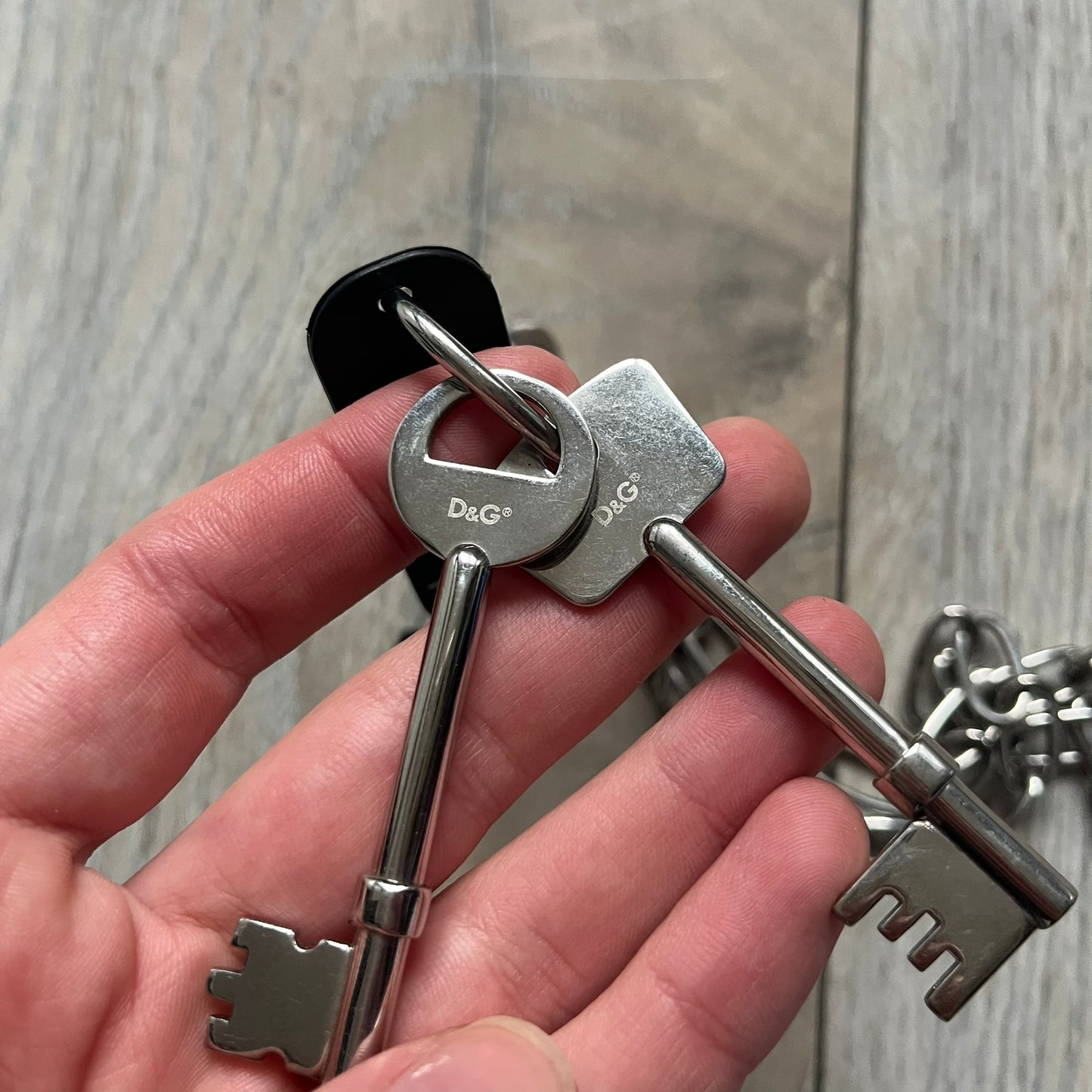 The Dolce &amp; Gabbana Room 13 Carabiner Necklace (DJ0613) is a large, heavy silver-tone necklace featuring a thick paperclip chain with oversized links. At the end of the chain is a large lobster clasp engraved with D&amp;G markings, connected to a branded carabiner. Hanging from the carabiner are two different keys with small D&amp;G logos and a black key identifier with a silver-tone plate engraved with “D&amp;G Dolce &amp; Gabbana Room 13.” The necklace comes with a large silver Dolce &amp; Gabbana dus