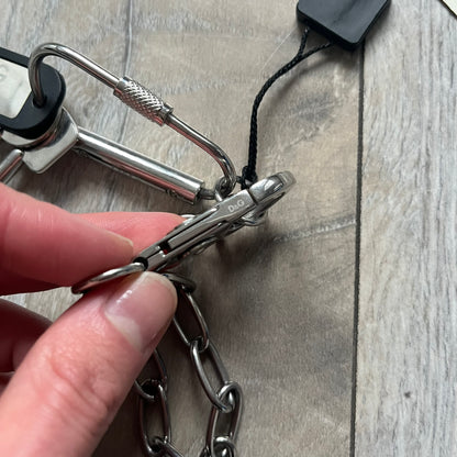 The Dolce &amp; Gabbana Room 13 Carabiner Necklace (DJ0613) is a large, heavy silver-tone necklace featuring a thick paperclip chain with oversized links. At the end of the chain is a large lobster clasp engraved with D&amp;G markings, connected to a branded carabiner. Hanging from the carabiner are two different keys with small D&amp;G logos and a black key identifier with a silver-tone plate engraved with “D&amp;G Dolce &amp; Gabbana Room 13.” The necklace comes with a large silver Dolce &amp; Gabbana dus