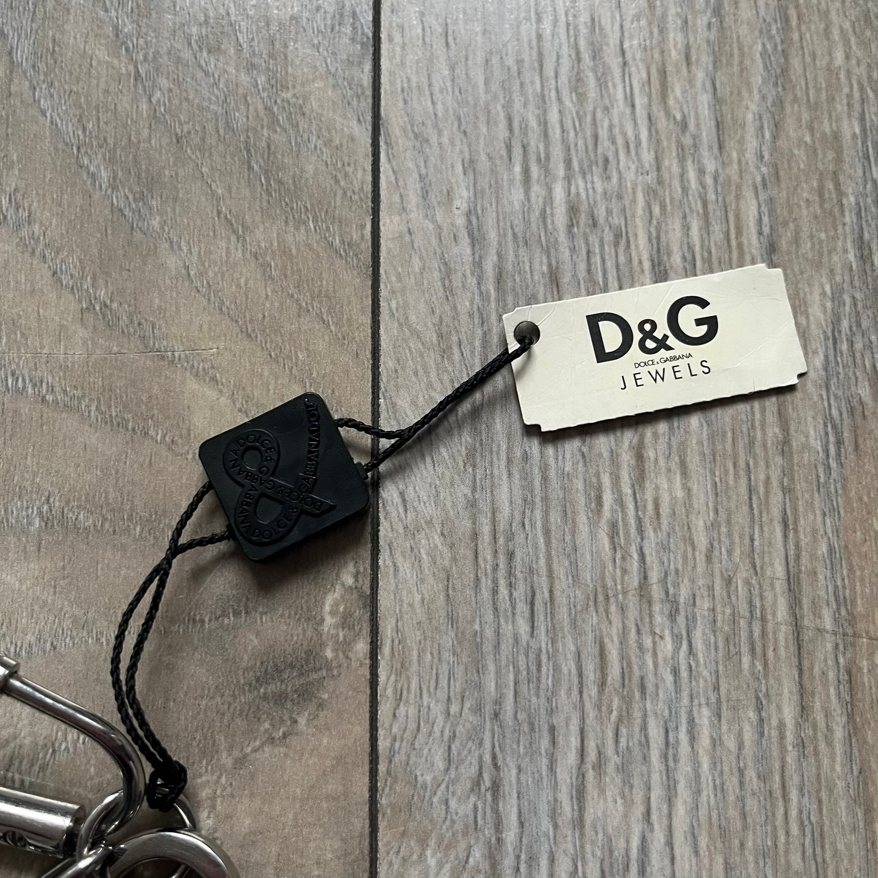 The Dolce &amp; Gabbana Room 13 Carabiner Necklace (DJ0613) is a large, heavy silver-tone necklace featuring a thick paperclip chain with oversized links. At the end of the chain is a large lobster clasp engraved with D&amp;G markings, connected to a branded carabiner. Hanging from the carabiner are two different keys with small D&amp;G logos and a black key identifier with a silver-tone plate engraved with “D&amp;G Dolce &amp; Gabbana Room 13.” The necklace comes with a large silver Dolce &amp; Gabbana dus