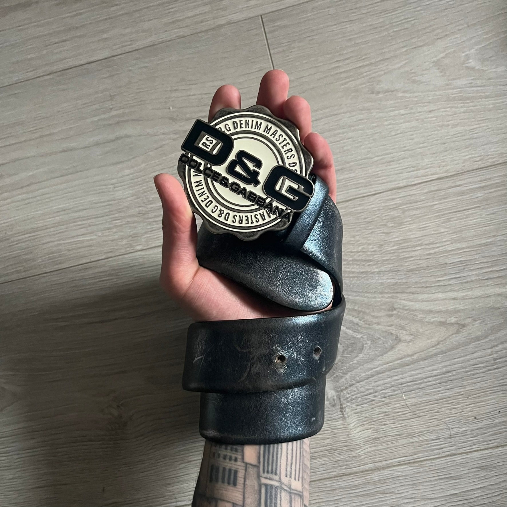 Dolce &amp; Gabbana Round Stamp Belt with a circular, wavy-edged buckle in a metallic finish. The buckle features a beige center with a bold black D&amp;G logo and “Dolce &amp; Gabbana” text beneath. Belt strap is glittery metallic blue with a black inner lining.