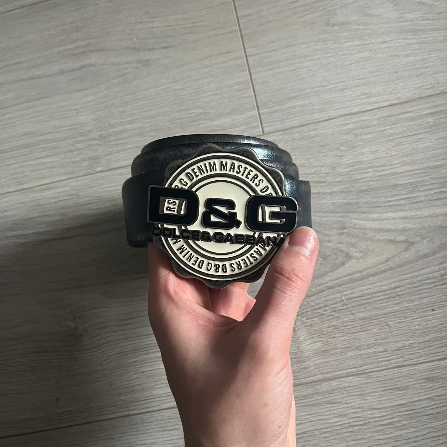 Dolce &amp; Gabbana Round Stamp Belt with a circular, wavy-edged buckle in a metallic finish. The buckle features a beige center with a bold black D&amp;G logo and “Dolce &amp; Gabbana” text beneath. Belt strap is glittery metallic blue with a black inner lining.
