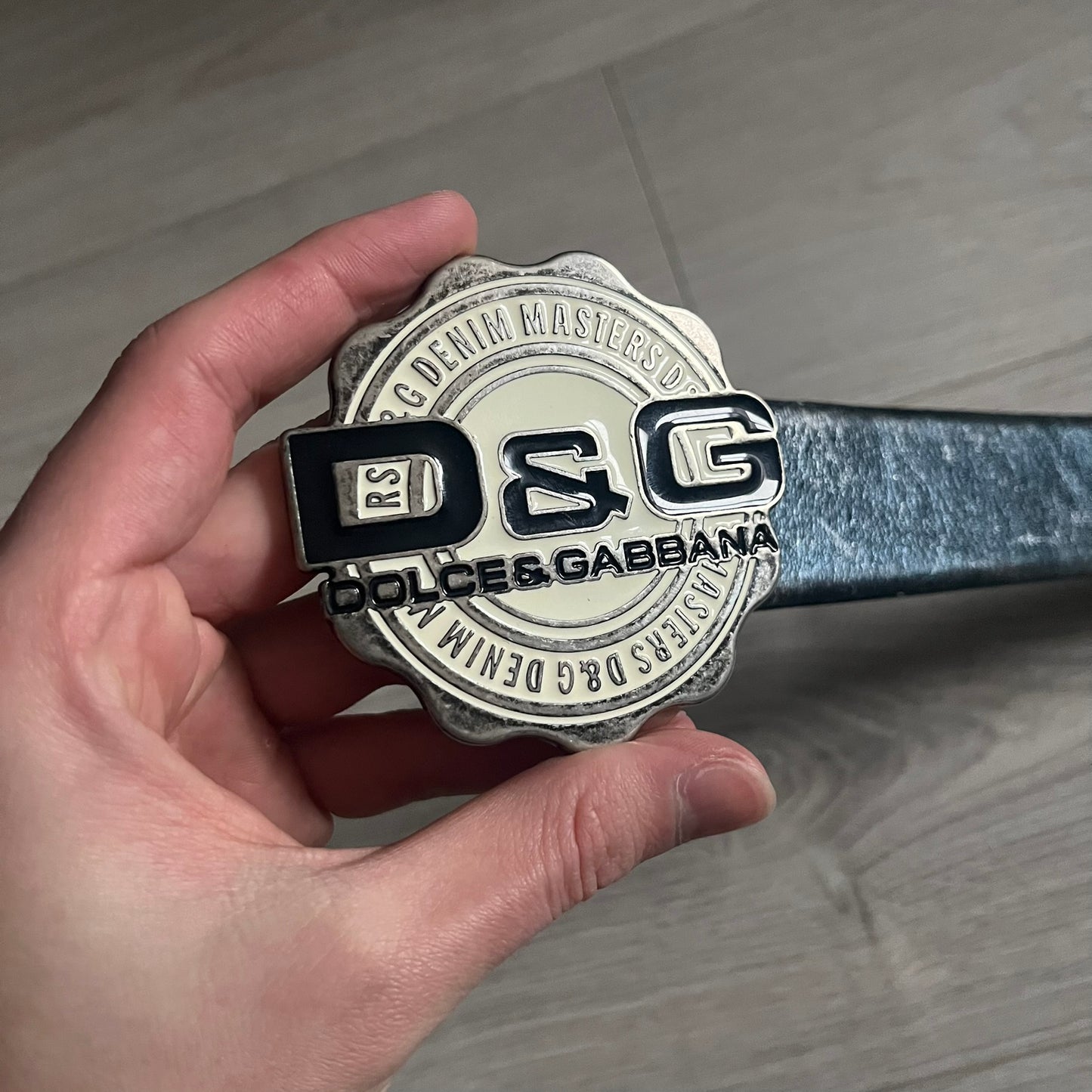 Dolce &amp; Gabbana Round Stamp Belt with a circular, wavy-edged buckle in a metallic finish. The buckle features a beige center with a bold black D&amp;G logo and “Dolce &amp; Gabbana” text beneath. Belt strap is glittery metallic blue with a black inner lining.
