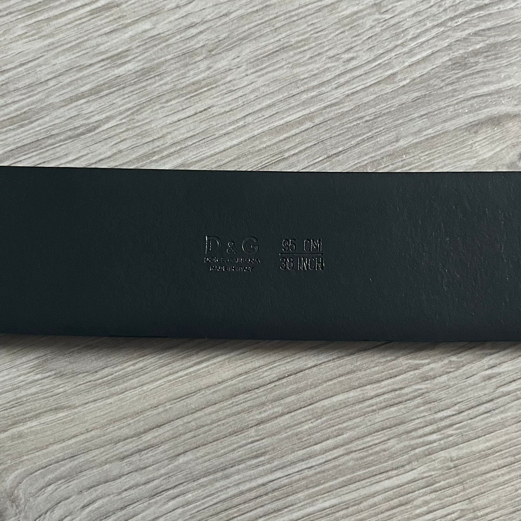 Dolce &amp; Gabbana Round Stamp Belt with a circular, wavy-edged buckle in a metallic finish. The buckle features a beige center with a bold black D&amp;G logo and “Dolce &amp; Gabbana” text beneath. Belt strap is glittery metallic blue with a black inner lining.