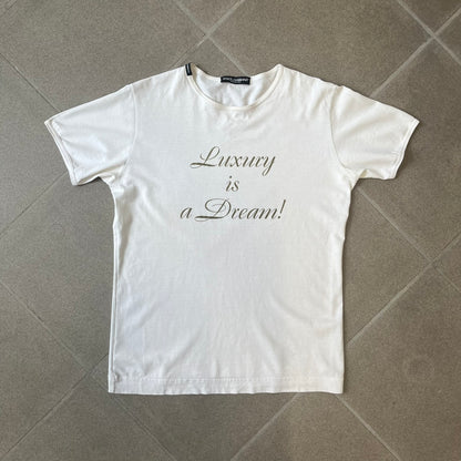 Dolce & Gabbana SS05 "Luxury Is a Dream!" White Tee features a clean white cotton design with dark gold handwritten-style text across the front reading “Luxury Is a Dream!”. The graphic uses a decorative quill-like font for a refined luxury aesthetic. Made in Italy, the tee includes two black Dolce & Gabbana tags, one at the neck and one on the collar.
