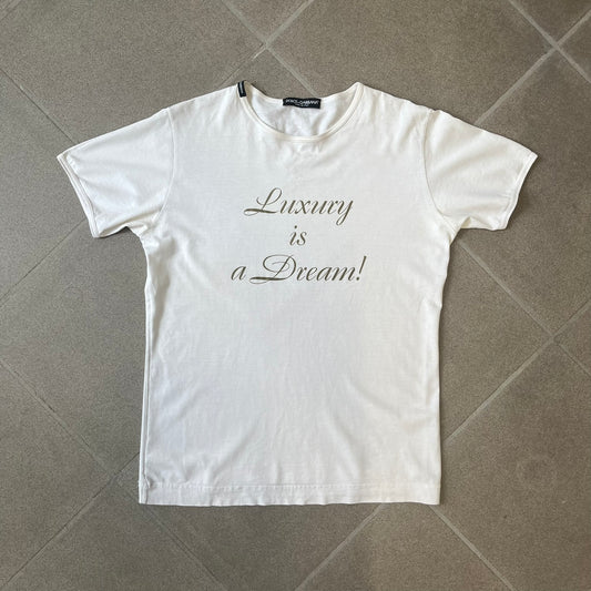 Dolce & Gabbana SS05 "Luxury Is a Dream!" White Tee features a clean white cotton design with dark gold handwritten-style text across the front reading “Luxury Is a Dream!”. The graphic uses a decorative quill-like font for a refined luxury aesthetic. Made in Italy, the tee includes two black Dolce & Gabbana tags, one at the neck and one on the collar.
