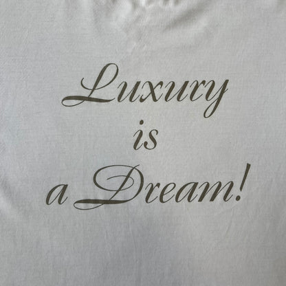 Dolce & Gabbana SS05 "Luxury Is a Dream!" White Tee features a clean white cotton design with dark gold handwritten-style text across the front reading “Luxury Is a Dream!”. The graphic uses a decorative quill-like font for a refined luxury aesthetic. Made in Italy, the tee includes two black Dolce & Gabbana tags, one at the neck and one on the collar.
