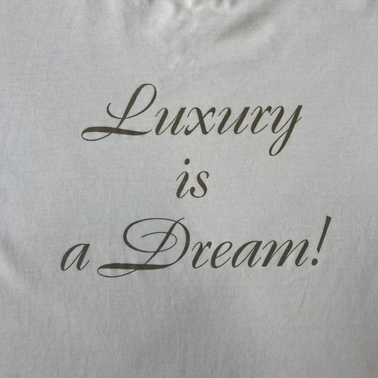 Dolce & Gabbana SS05 "Luxury Is a Dream!" White Tee features a clean white cotton design with dark gold handwritten-style text across the front reading “Luxury Is a Dream!”. The graphic uses a decorative quill-like font for a refined luxury aesthetic. Made in Italy, the tee includes two black Dolce & Gabbana tags, one at the neck and one on the collar.
