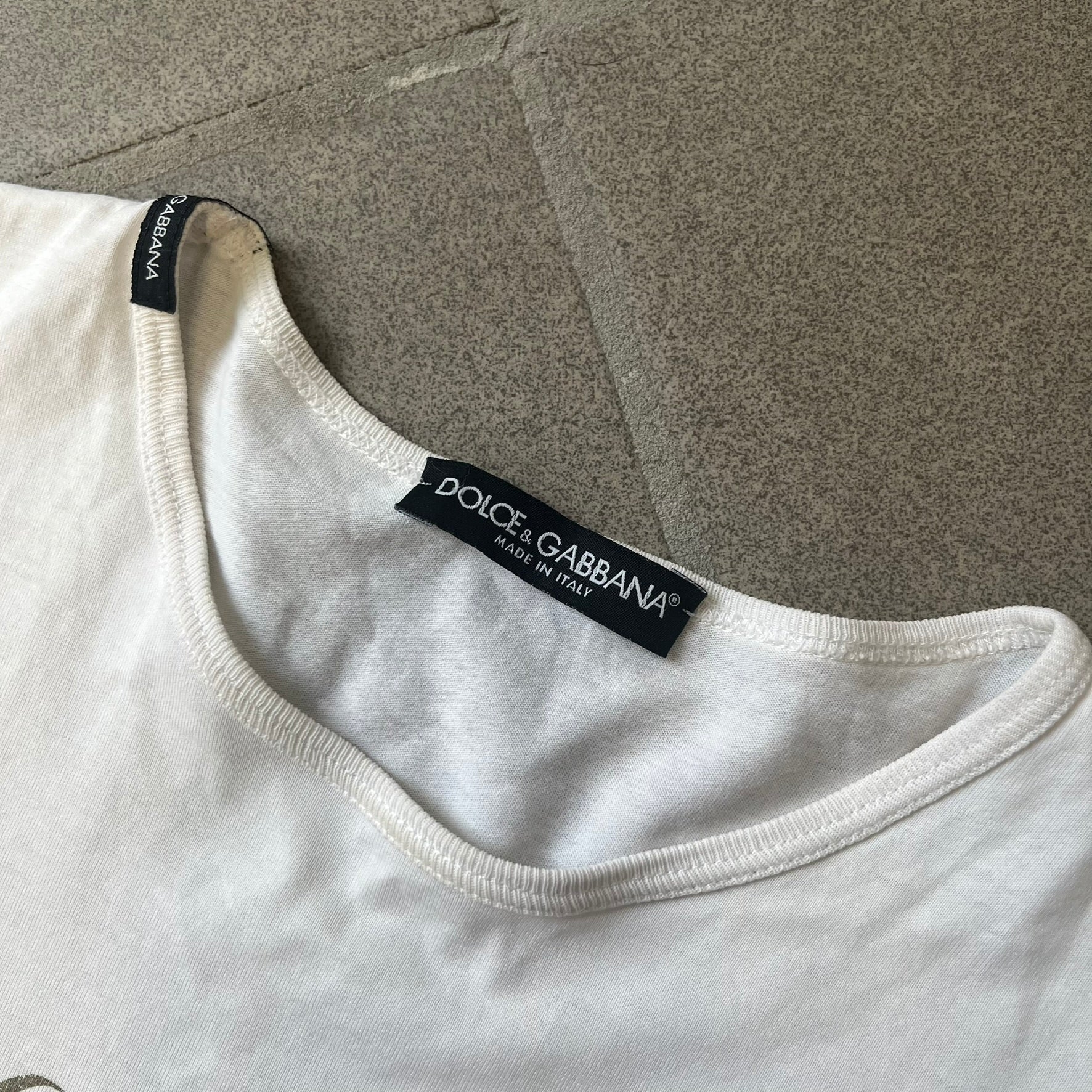 Dolce & Gabbana SS05 "Luxury Is a Dream!" White Tee features a clean white cotton design with dark gold handwritten-style text across the front reading “Luxury Is a Dream!”. The graphic uses a decorative quill-like font for a refined luxury aesthetic. Made in Italy, the tee includes two black Dolce & Gabbana tags, one at the neck and one on the collar.
