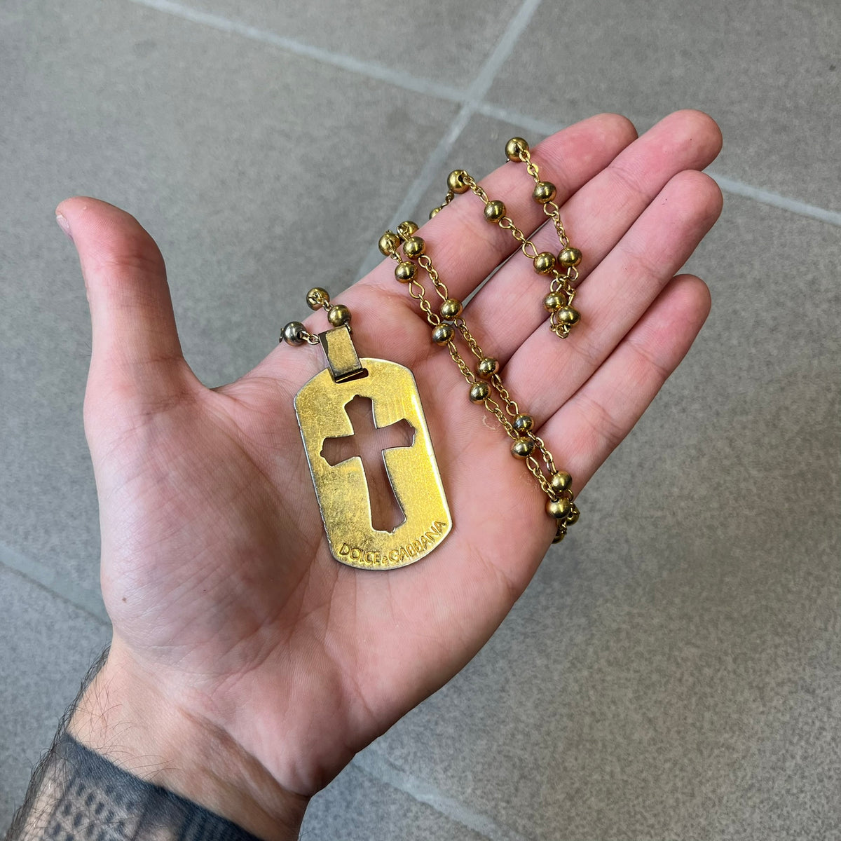 Dolce & Gabbana SS2003 Golden Cross Dog Tag Necklace features a curved-edge dog tag pendant with a cutout Latin cross in the center and engraved "Dolce & Gabbana" script text below it. It comes on a thin chain set with fixed beaded accents at regular intervals.