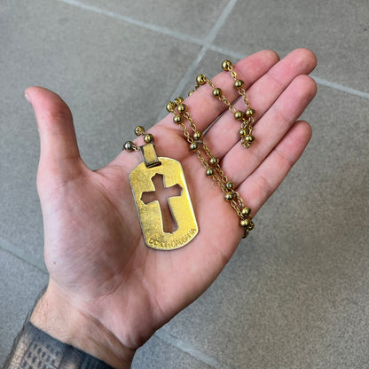 Dolce & Gabbana SS2003 Golden Cross Dog Tag Necklace features a curved-edge dog tag pendant with a cutout Latin cross in the center and engraved "Dolce & Gabbana" script text below it. It comes on a thin chain set with fixed beaded accents at regular intervals.
